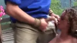 Blonde Lockenkopf-Frau kniet vor Mann in blauem Shirt und lutscht gierig seinen Schwanz auf sonniger Holzterrasse.